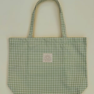 May include: A light green and white gingham tote bag with two matching fabric handles. A small square label is centered on the front, featuring a sun icon and the text SUN FRECKLED STUDIO printed in a dark brown color on a light background.