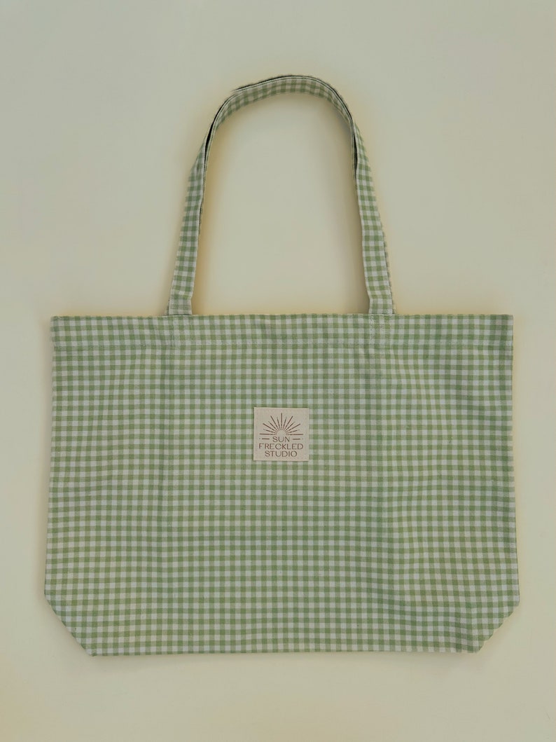 May include: A light green and white gingham tote bag with two matching fabric handles. A small square label is centered on the front, featuring a sun icon and the text SUN FRECKLED STUDIO printed in a dark brown color on a light background.