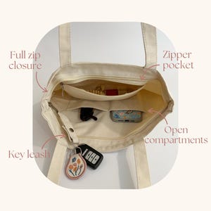 May include: A cream-colored canvas tote bag with an open top, featuring a full zip closure, an interior zipper pocket, two open compartments, and a key leash. The bag contains a set of car keys with a floral embroidered keychain attached.