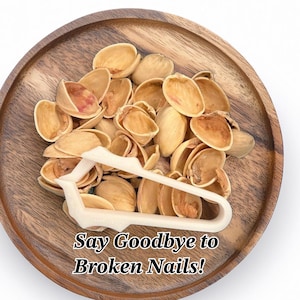 May include: A white plastic pistachio nutcracker on a wooden plate with a pile of pistachio shells. The text "Say Goodbye to Broken Nails!" is written below the nutcracker.
