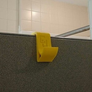 May include: A yellow plastic hook with a square design on the front is mounted on a gray wall.