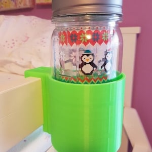 May include: A green plastic holder for a glass jar with a silver lid. The jar has a penguin design with a red and white pattern.