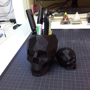 May include: A black skull-shaped desk organizer with a geometric design. The organizer is filled with pens and pencils. There is a smaller black skull-shaped object next to the organizer.