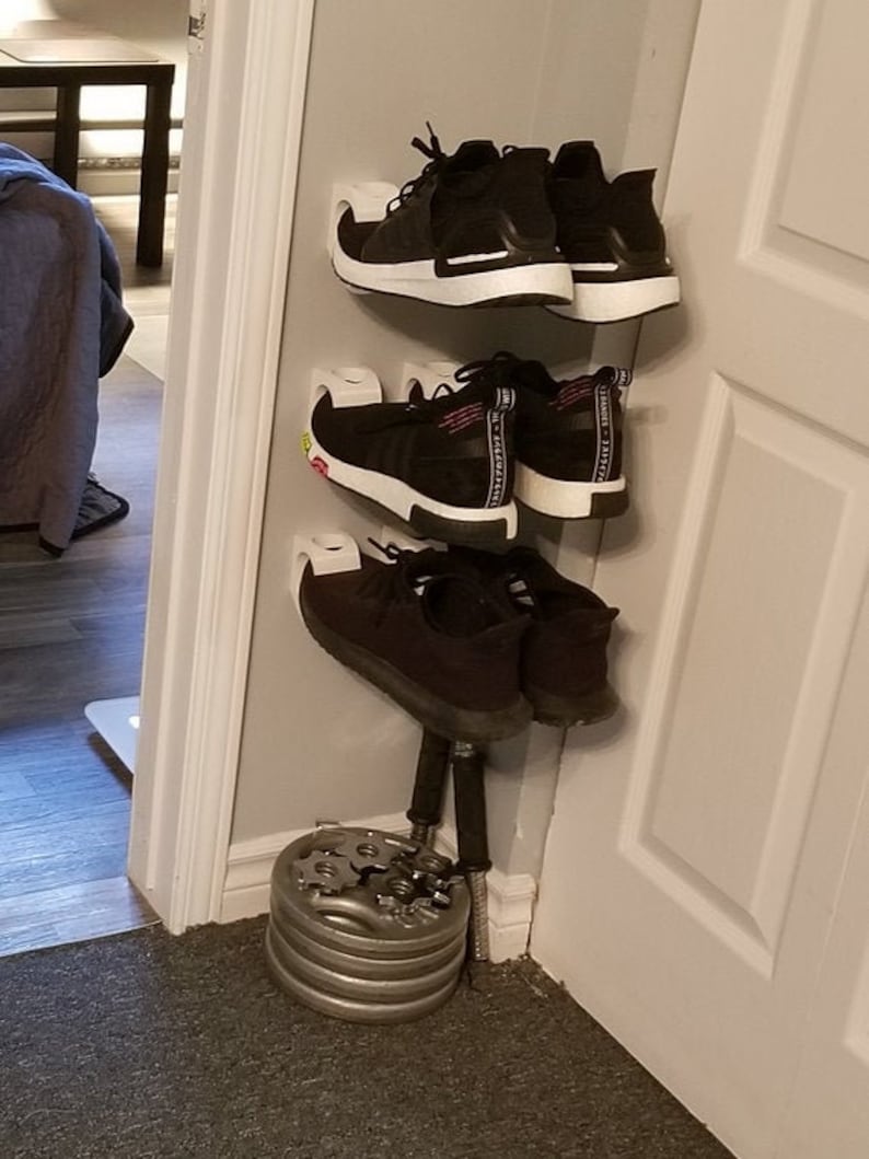 May include: A white shoe rack with three shelves holding black sneakers. The rack is mounted on a white wall in a corner. A set of silver weight plates are stacked on the floor below the rack.