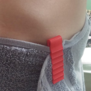 Könnte beinhalten: Ein roter Plastik-Handtuchclip mit strukturierter Oberfläche. Der Clip ist an einem grauen Handtuch befestigt.