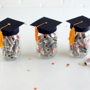 Peut inclure: Trois bocaux en verre remplis de billets de dollars enroulés, chacun surmonté d'un bonnet de graduation noir avec un pompon doré. Les bocaux sont sur une surface blanche avec des confettis orange et noirs.