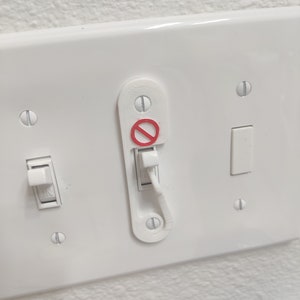 May include: A white light switch plate with two toggle switches and a single outlet. The outlet has a white plastic cover with a red circle and a diagonal line through it, indicating that it is not to be used.