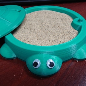 May include: A green plastic turtle sandbox with a lid. The sandbox is filled with sand and has googly eyes.