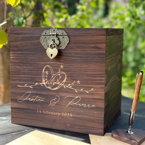 May include: A dark brown wooden box with a decorative clasp and heart-shaped lock. The box is engraved with the names "Justine & Pierre" and the date "14 february 2023". A wooden pen and heart-shaped stand are next to the box.