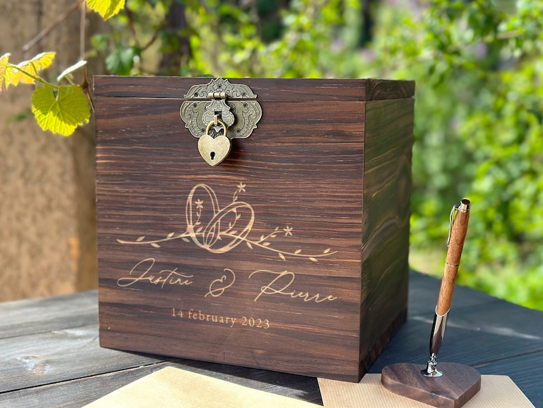 May include: A dark brown wooden box with a decorative clasp and heart-shaped lock. The box is engraved with the names "Justine & Pierre" and the date "14 february 2023". A wooden pen and heart-shaped stand are next to the box.