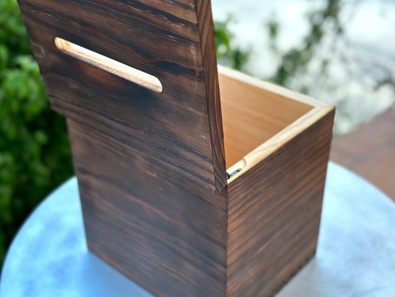 May include: A handcrafted wooden box with a hinged lid and a rectangular handle cutout. The box is made of dark stained wood, showcasing the natural grain. The interior is a lighter wood color.