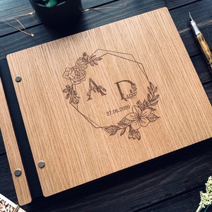 May include: A wooden guest book with a black leather spine and metal accents. The cover features a laser-engraved design of two initials "A" and "D" surrounded by a floral wreath and a geometric frame. The date "27.06.2020" is also engraved on the cover.