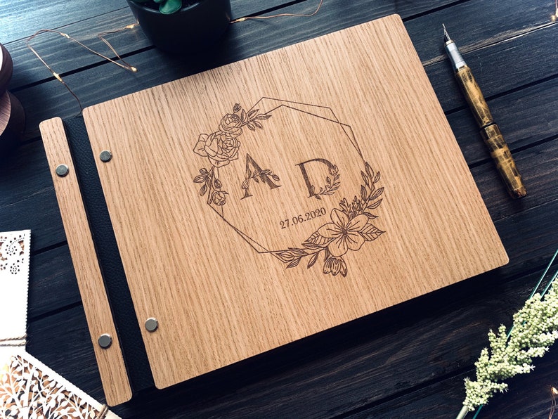 May include: A wooden guest book with a black leather spine and metal accents. The cover features a laser-engraved design of two initials "A" and "D" surrounded by a floral wreath and a geometric frame. The date "27.06.2020" is also engraved on the cover.