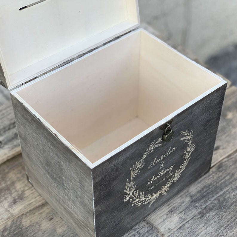 May include: A gray wooden box with a white interior and a hinged lid. The box has a small metal lock and keyhole. The front of the box is decorated with a wreath of leaves and the text "Aurelia & Anthony 12 September 2020".