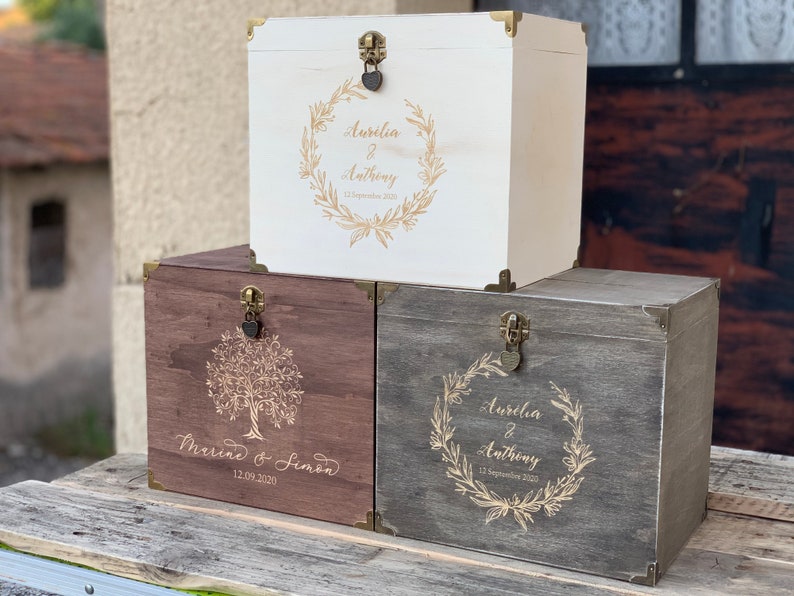 K&ouml;nnte beinhalten: Drei Holzkisten mit gravierten Designs und goldenen Akzenten. Die obere Kiste ist wei&szlig; mit einem Kranzdesign und dem Text "Aurelia & Anthony 13. September 2020". Die mittlere Kiste ist braun mit einem Baumdesign und dem Text "Marine & Simon 12.09.2020". Die untere Kiste ist grau mit einem Kranzdesign und dem Text "Aurelia & Anthony 13. September 2020".