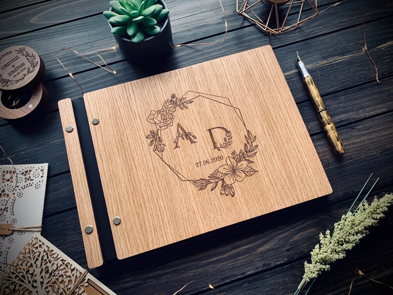 May include: A wooden guest book with a black leather binding. The cover is engraved with a floral design, the initials "AD", and the date "27.06.2020".