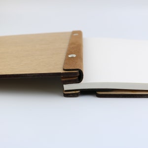 May include: A wooden journal with a brown leather spine and metal rivets. The journal is open to reveal blank white pages.