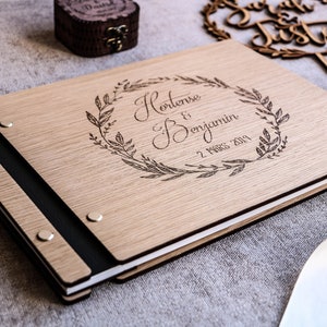 May include: A rectangular wooden photo album with a light brown wood grain finish. The cover features a laser-engraved laurel wreath surrounding the text Hortense & Benjamin and the date 2 MARS 2019. It has a black spine secured by four silver metal rivets.