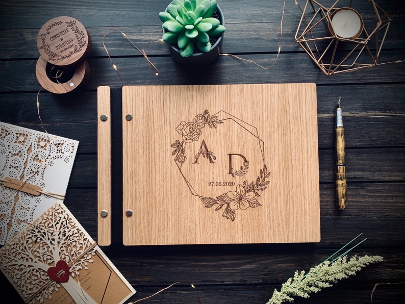 May include: A wooden guest book with a floral design and the initials "A D" engraved on the front. The date "27.06.2020" is also engraved on the book.