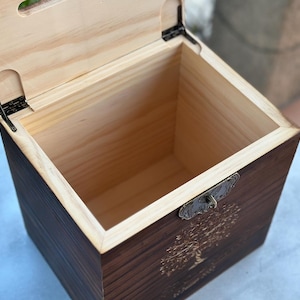 May include: Open wooden box with a hinged lid. The box is made of dark stained wood with a lighter wood interior. A decorative metal clasp and a tree design are visible on the front.