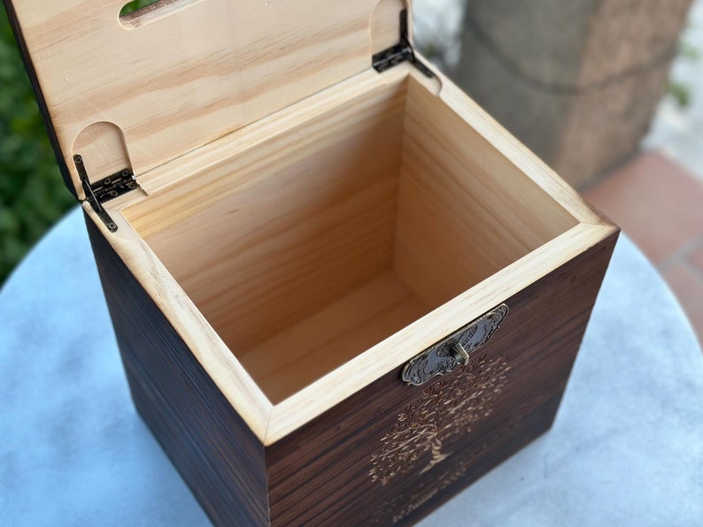 May include: Open wooden box with a hinged lid. The box is made of dark stained wood with a lighter wood interior. A decorative metal clasp and a tree design are visible on the front.