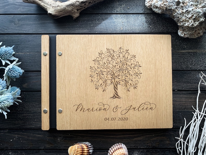 May include: A wooden guest book with a black spine and a laser engraved tree design. The text "Marion & Julien 04.07.2020" is engraved below the tree.