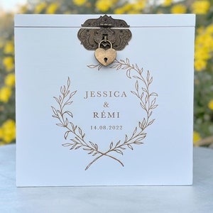 May include: A white wooden box with a decorative bronze clasp and heart-shaped lock. The box features a gold-colored wreath design with the names "JESSICA & R&Eacute;MI" and the date "14.08.2022" printed on the front. The box is likely for a wedding or special event.