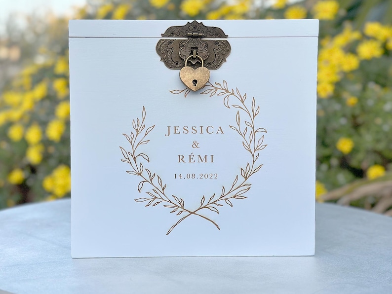 May include: A white wooden box with a decorative bronze clasp and heart-shaped lock. The box features a gold-colored wreath design with the names "JESSICA & R&Eacute;MI" and the date "14.08.2022" printed on the front. The box is likely for a wedding or special event.