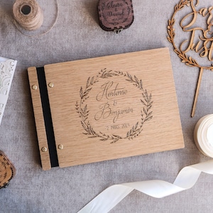 May include: A rectangular wooden photo album with a black spine and metal binding screws. The cover features a laser-engraved laurel wreath surrounding the text Hortense & Benjamin and 2 MARS 2019. The album is surrounded by wedding stationery, wooden boxes, and a cake topper.