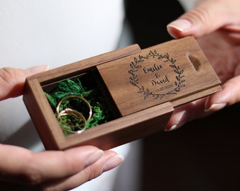 May include: A wooden ring box with a sliding lid. The lid is engraved with a wreath of leaves and the text "Emilie & David 11.07.2020". The box is open and contains two gold rings on a bed of green moss.