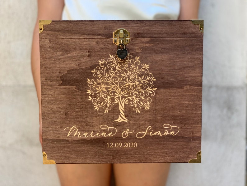 K&ouml;nnte beinhalten: Eine Holzkiste mit einem goldenen Baumdesign und den Namen "Marine & Simon" sowie dem Datum "12.09.2020" auf der Vorderseite graviert. Die Kiste hat ein goldenes Schloss und ein Schl&uuml;sselloch.