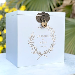 May include: A white wooden box with a decorative lock and a heart-shaped key. The box features the names "JESSICA & R&Eacute;MI" and the date "14.08.2022" within a gold-colored wreath design. The box is likely used for a wedding.