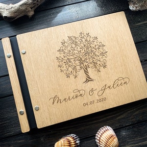 May include: A wooden photo album with a black spine and metal accents. The front cover features a laser-engraved design of a tree with intricate branches and the text "Mariam & Julien 04.07.2020".