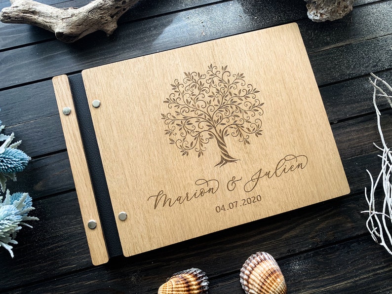May include: A wooden photo album with a black spine and metal accents. The front cover features a laser-engraved design of a tree with intricate branches and the text "Mariam & Julien 04.07.2020".