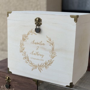 K&ouml;nnte beinhalten: Wei&szlig;e Holzkiste mit goldenem Schloss und Scharnieren. Die Kiste ist mit einem goldenen Blumengirlanden und dem Text "Aurelia & Anthony 12 Septembre 2020" verziert.