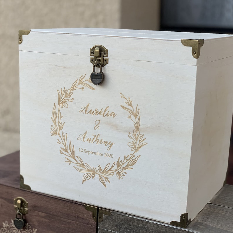 K&ouml;nnte beinhalten: Wei&szlig;e Holzkiste mit goldenem Schloss und Scharnieren. Die Kiste ist mit einem goldenen Blumengirlanden und dem Text "Aurelia & Anthony 12 Septembre 2020" verziert.