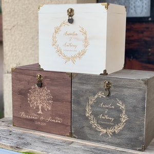 Peut inclure: Trois boîtes en bois avec des motifs gravés et des accents dorés. La boîte du haut est blanche avec un motif de couronne et le texte "Aurelia & Anthony 13 septembre 2020". La boîte du milieu est brune avec un motif d'arbre et le texte "Marine & Simon 12.09.2020". La boîte du bas est grise avec un motif de couronne et le texte "Aurelia & Anthony 13 septembre 2020".