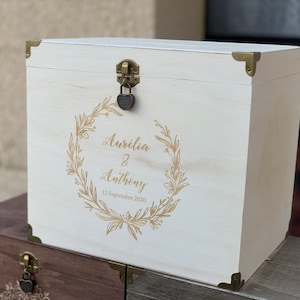 May include: White wooden box with a gold lock and hinges. The box is decorated with a gold floral wreath and the text "Aurelia & Anthony 12 Septembre 2020".