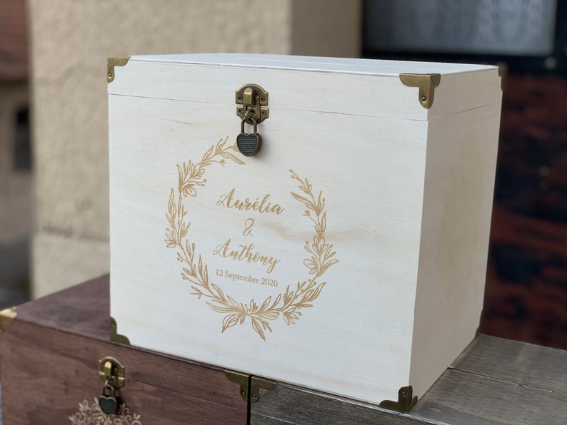 May include: White wooden box with a gold lock and hinges. The box is decorated with a gold floral wreath and the text "Aurelia & Anthony 12 Septembre 2020".