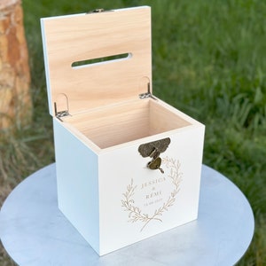 May include: A white wooden box with a hinged lid and a slot on top. The box has a decorative lock and a gold-colored floral design with the names "Jessica & Remi" and the date "11.06.2022" printed on the front. The box is sitting on a round, light-colored surface.