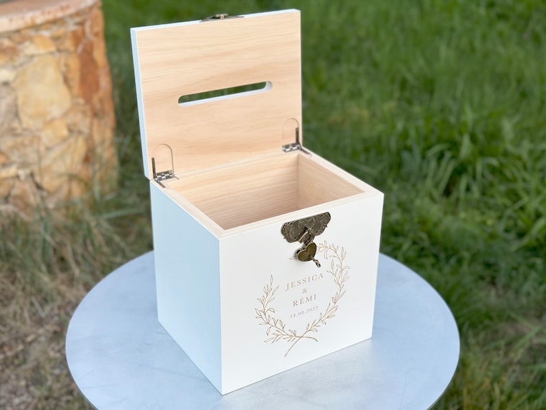 May include: A white wooden box with a hinged lid and a slot on top. The box has a decorative lock and a gold-colored floral design with the names "Jessica & Remi" and the date "11.06.2022" printed on the front. The box is sitting on a round, light-colored surface.