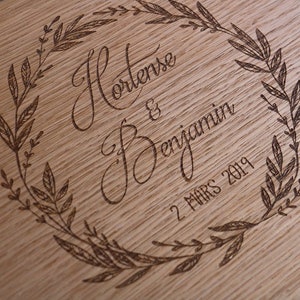 May include: A light brown wooden board featuring a laser-engraved design of a circular leaf wreath. Inside the wreath, the names Hortense & Benjamin are written in a cursive script, followed by the date 2 MARS 2019 in a simple font.