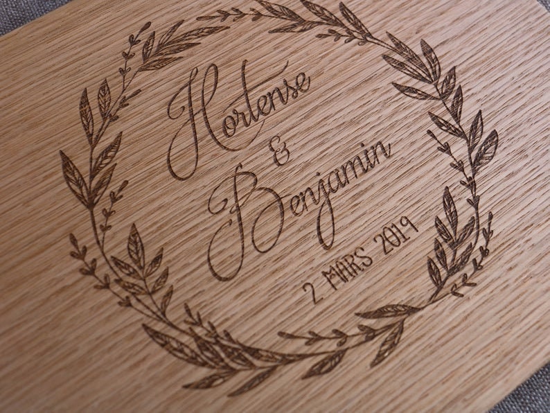 May include: A light brown wooden board featuring a laser-engraved design of a circular leaf wreath. Inside the wreath, the names Hortense & Benjamin are written in a cursive script, followed by the date 2 MARS 2019 in a simple font.