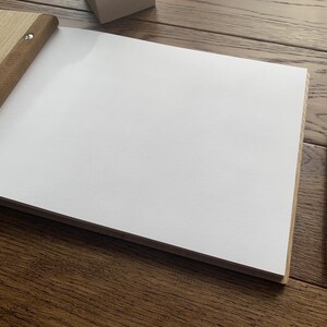 May include: A blank, white page in a brown leather-bound journal with a wooden cover. A gold pen is lying on a wooden surface next to the journal.
