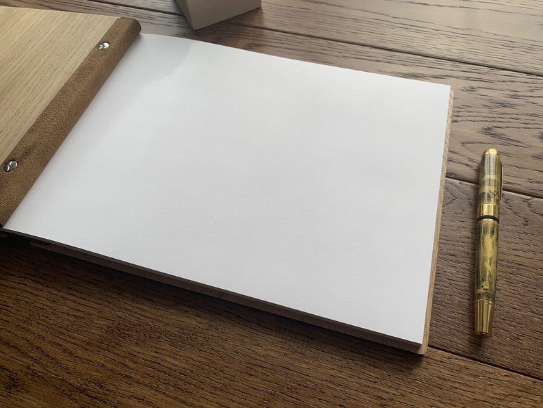 May include: A blank, white page in a brown leather-bound journal with a wooden cover. A gold pen is lying on a wooden surface next to the journal.