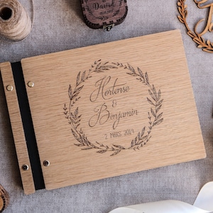 May include: A rectangular wooden photo album with a black spine and metal binding screws. The front cover features a laser-engraved laurel wreath surrounding the names Hortense & Benjamin and the date 2 MARS 2019 in a script and block font.