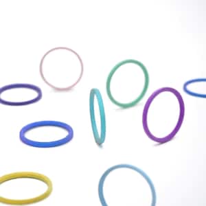 Può includere: Una collezione di anelli sottili e colorati in varie tonalità di blu, viola, verde, rosa e giallo. Gli anelli sono sparsi su uno sfondo bianco, creando una composizione visivamente accattivante. Gli anelli sembrano essere fatti di un materiale flessibile.