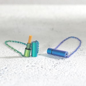Könnte beinhalten: Zwei kleine, zylindrische Metallbehälter mit Kettenanhängern. Einer ist grün und goldfarben, der andere blau und lila. Der grün-goldene Behälter hat einen kleinen, hellbraunen Gegenstand, der daraus herausragt.