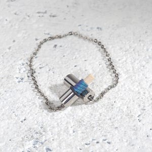 Könnte beinhalten: Ein silberfarbenes Armband mit einem kreuzförmigen Anhänger. Der Anhänger besteht aus zwei zylindrischen Metallteilen, die mit blauem und violettem Faden umwickelt sind, und einem kleinen Holzelement. Das Armband hat eine feine Kette.