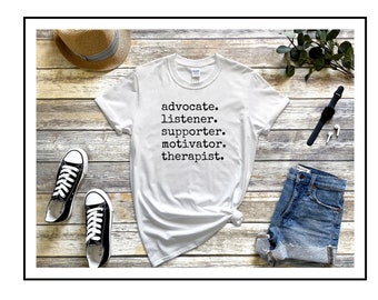 以下が含まれることがあります： 黒い文字で「advocate. listener. supporter. motivator. therapist.」とプリントされた白いTシャツ